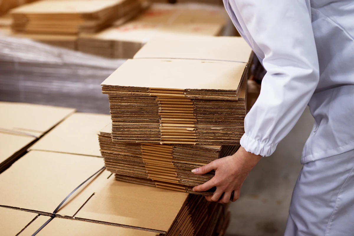 close up young female worker picking up stacks folded cardboard boxes from bigger stack factory storage room โรงงานผลิตกล่องกระดาษลูกฟูก,โรงงานผลิตกล่อง,รับผลิตกล่องกระดาษลูกฟูก,กล่องกระดาษลูกฟูก,กล่องสั่งผลิต,กล่องพิมพ์ลาย,กล่องคุณภาพดี